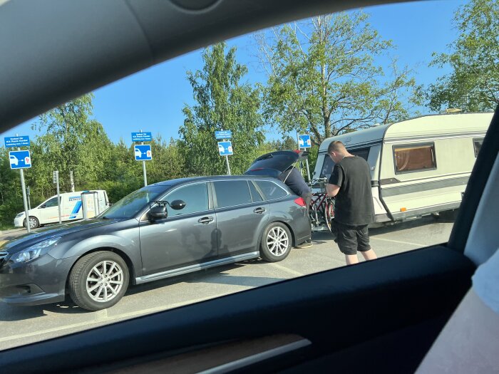 En parkering med person, grå bil, husvagn och blå skyltar under solig himmel.