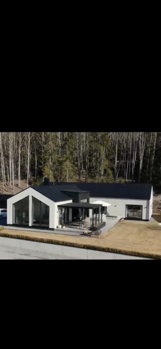 Ett modernt hus med stora fönster, platt tak, och omgivet av skog nära en väg.