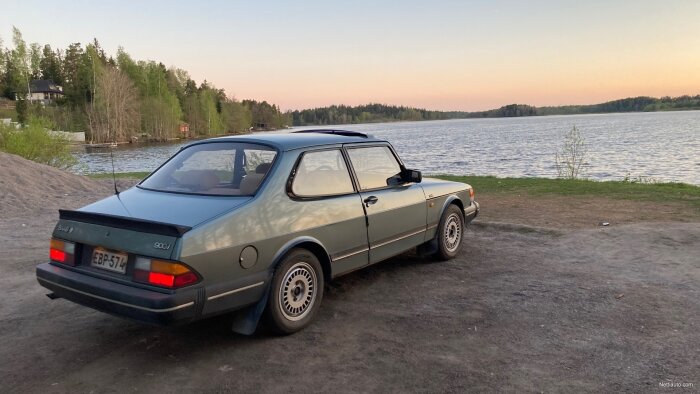 En klassisk Saab 900 bil parkerad vid en lugn sjö vid skymningen. Träd och hus i bakgrunden.