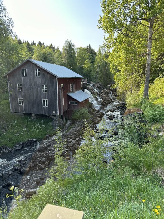 En träbyggnad med röda detaljer intill en forsrännande ström omgiven av grönska och skog.