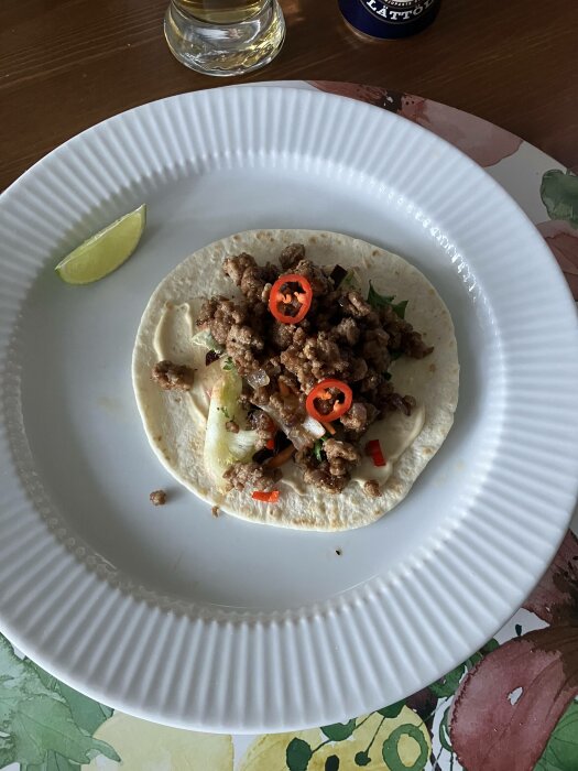 En taco med köttfärs, koriander och skivad chili. Limeklyfta och ölglas i bakgrunden. Mat på tallrik.