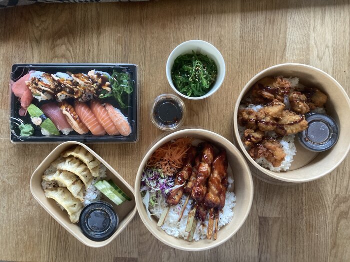 Olika japanska rätter inklusive sushi, dumplings, sallader och kycklingspett serverade i behållare på ett träbord.