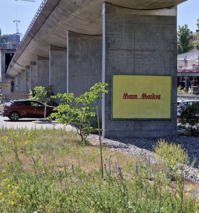 Stadsmiljö med betongbro, skylt med texten "Mmmm...Moskva", grönska, bil och byggnader i bakgrunden.