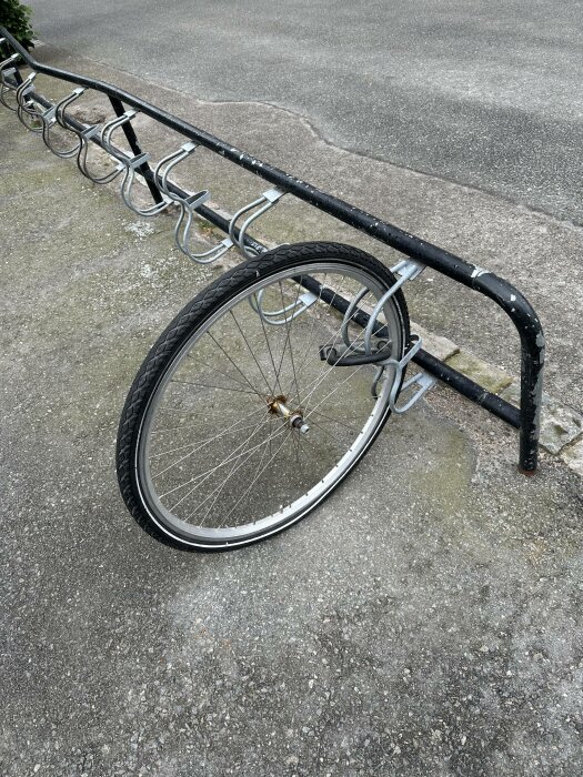 Framhjul på cykel låst vid ställning, resten av cykeln saknas. Grå asfalt, dagtid.