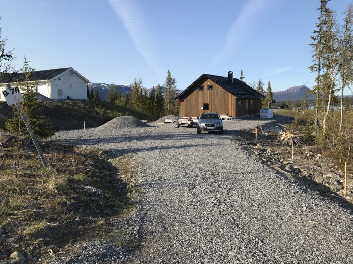 En bilfrämre uppfart till ett trähus, grushögar, skog, fjäll i bakgrunden, klar himmel.