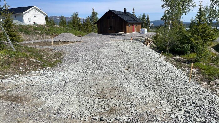 Grusväg leder till moderna hus, bergig terräng, skogsbakgrund, byggmaterial, utvecklad infrastruktur och natur.