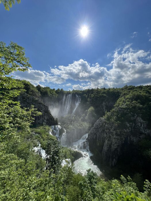 Solen skiner över ett frodigt vattenfall och gröna träd i en klippig terräng.