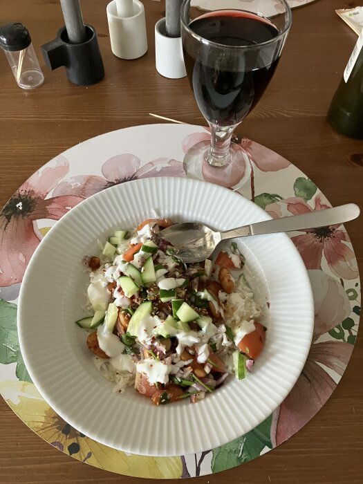 Maträtt med ris, grönsaker, röd sås, halloumi, gurka och rödvin, matbord med kryddkvarnar och servetter.