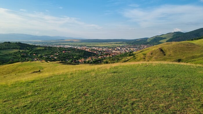 Lantlig vy, kullar, gröna ängar, stad i dal, berg i bakgrunden, klar himmel, naturskönt landskap.