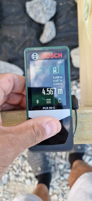 Hand håller en Bosch laseravståndsmätare som visar mätning: 4.567 meter. Bakgrunden är suddig, med sten och trä synligt.