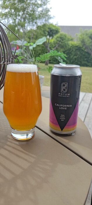 Ölglas fyllt med skummande öl, bredvid en burk märkt "California Love" från Potum Brewery. Uteservering och grönska i bakgrunden.