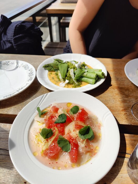 Färska rätter serverade utomhus: citrus carpaccio, gröna sparrisar, soligt bord, delvis tom tallrik, avslappnad atmosfär.