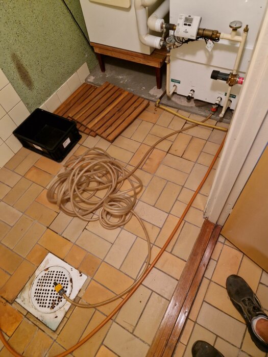 Ett badrumsgolv med slang, avloppsbrunn, husvattenmätare, och trädetaljer; tecken på arbete eller underhåll.