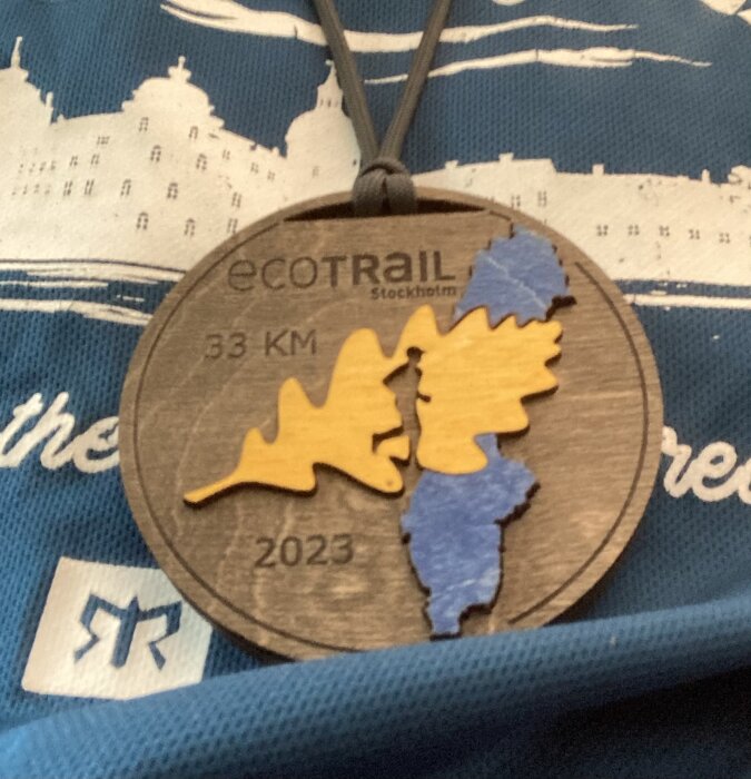 Medalj med texten "EcoTrail Stockholm 33 KM 2023", träd, och del av blå tröja.