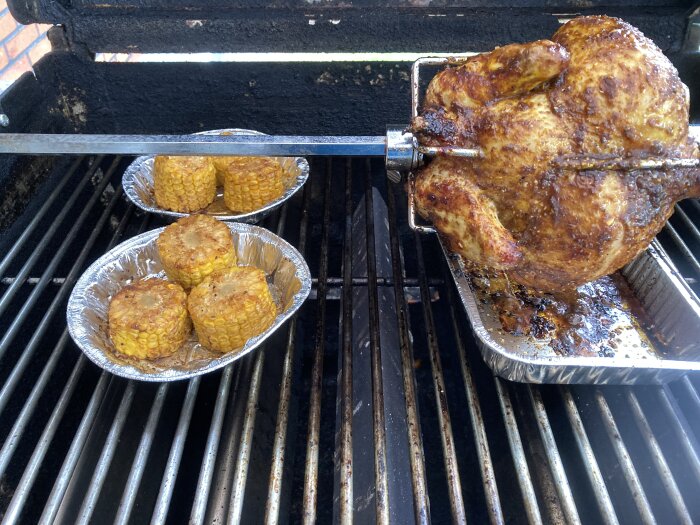 Hel kyckling roterar på spett över grill, majs i folieformar, matlagning utomhus.