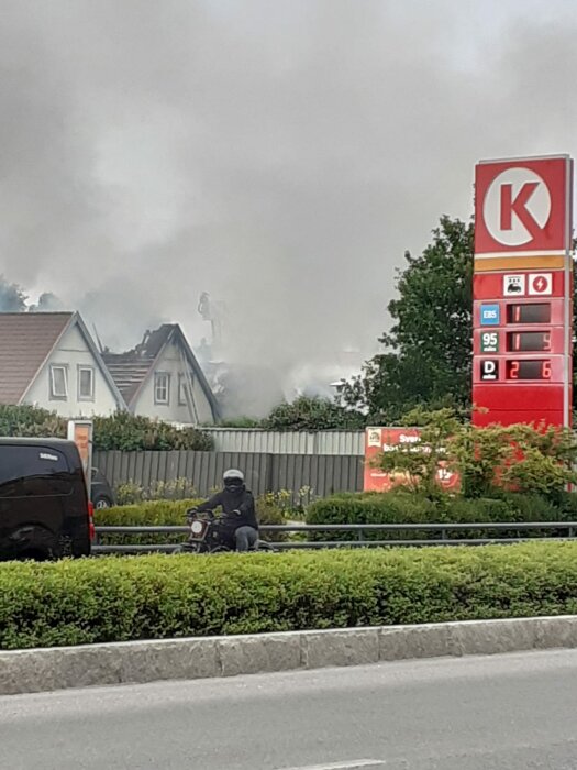 Rökutveckling bakom bensinstation, bil, person på motorcykel, träd, grå himmel.