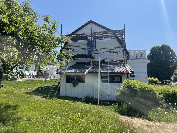 Hus med byggnadsställningar, grönt gräs, klar himmel, stege, solljus, träd, renovering pågår.