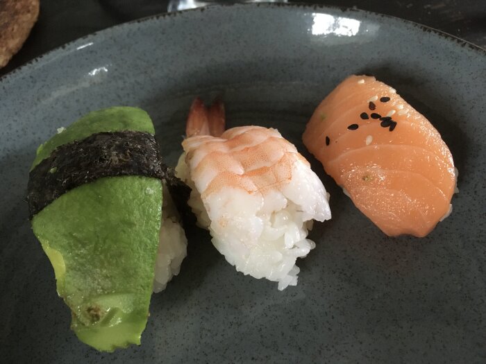 Tre sushi: avokado-nigiri, räka-nigiri, lax-nigiri på grå tallrik.