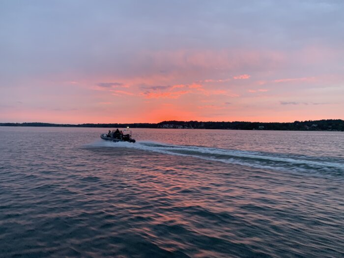 Motorbåt glider på vatten vid solnedgång med rosa himmel och silhuetter av träd.