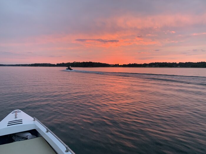 Solnedgång över lugnt vatten, båt framåt, väckor, färgerosa himmel, träd i silhuett.