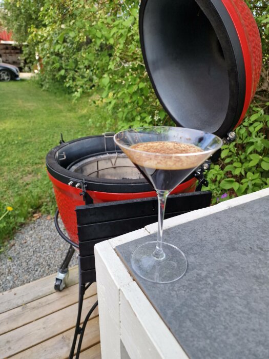 Martini glas med brun dryck ovanpå en väderbeständig yta, röd grill i bakgrunden, utomhusmiljö.