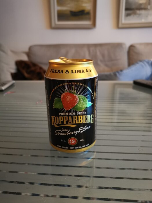 En burk Kopparberg cider med jordgubb och lime på ett glasbord, inomhus, suddig bakgrund, mysig atmosfär.