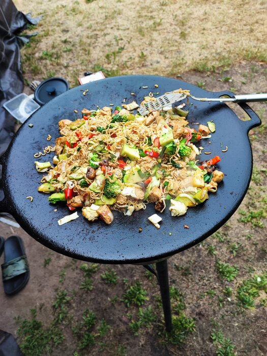 Stir-fry med grönsaker och nudlar på stor svart utomhuspanna. Matlagning utomhus, färgrik och aptitretande rätt.