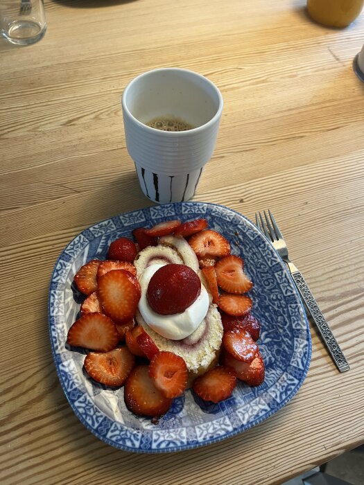 Ett fat med tårta, jordgubbar och grädde bredvid en kaffekopp på ett träbord.