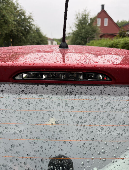 Röd bil med regndroppar, antenn och LED-ljus, mot suddig husbakgrund.