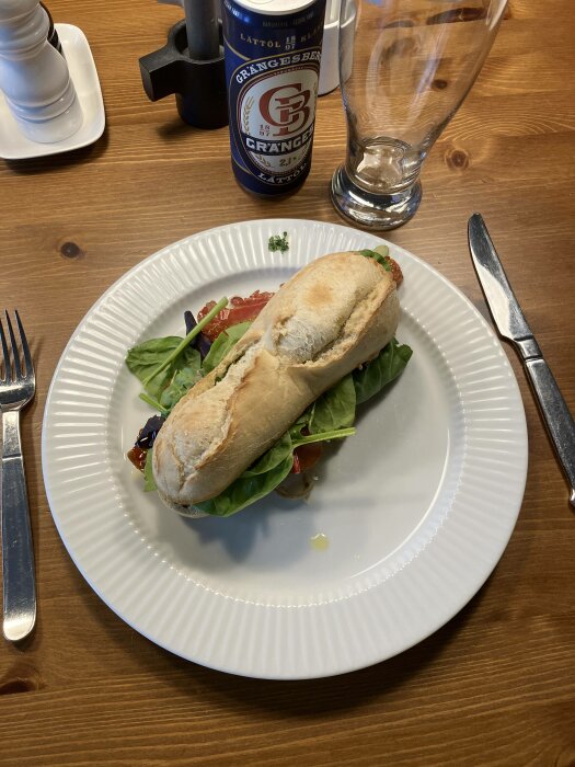 Ett smörgås med sallad på en vit tallrik, en ölburk, saltkar, pepparkvarn, glas och bestick på träbord.