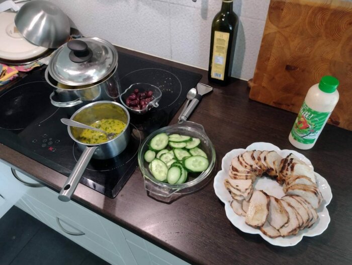 Köksbänk med matlagning: grytor, skivad gurka, bönor, grillat kött, dressing, skärbräda, spishäll.