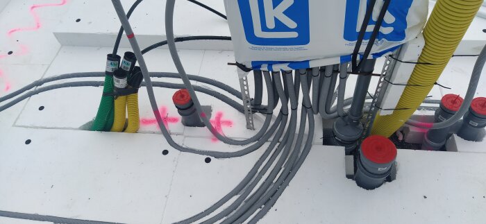 Byggarbetsplats med isoleringsmaterial, olika kablar och rör, märkning på golvet, samt en produkt med LK-logotyp.