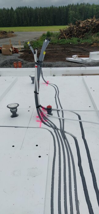 Kabelinstallation på tak med kondukt, isolering, och el-komponenter; byggarbetsplats i bakgrunden med grönska.