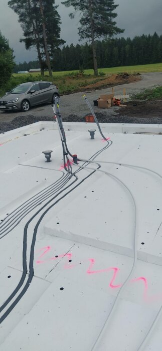 Vit takkonstruktion med svarta och grå rör, rosa markeringar, bil och träd i bakgrunden under molnig himmel.