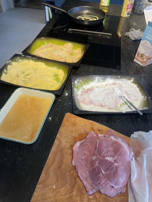 Kök med förberedelser för panering och stekning av kött. Ingredienser och köksredskap synliga.