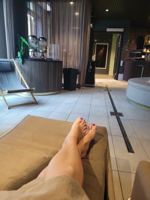 En person med blå nagellack avslappnad i en modern lounge med fåtöljer och dryckesbehållare i bakgrunden.