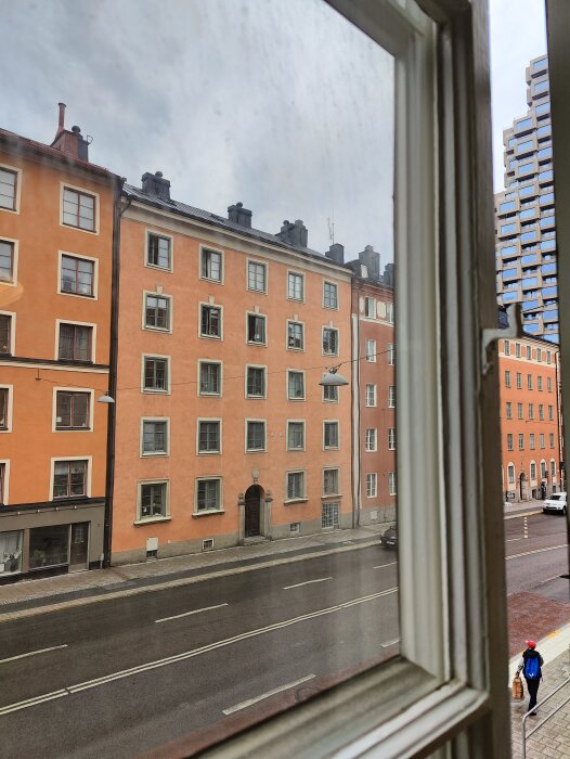 Bild från ett fönster på en stadsgata med orange byggnader, en gående person och regnig himmel.