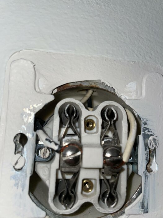 En närbild av en omonterad elektrisk uttagskontakt med synliga ledningar och anslutningar, omgiven av vit vägg.