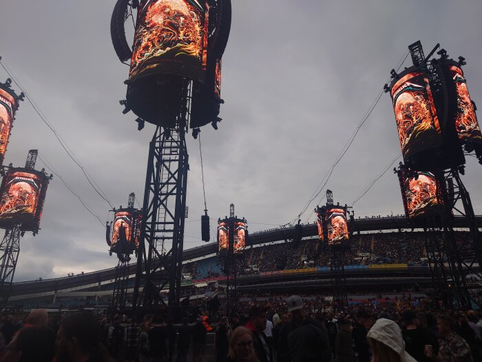 Stor konsert med publik, mörka moln, flera skärmar med flammande motiv, stor stadionatmosfär.