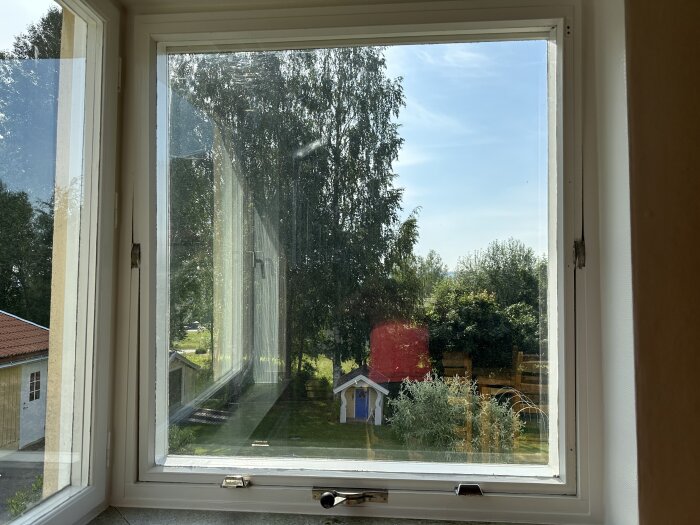Utsikt genom ett öppet fönster, träd, gräs, rött förråd, blå lekstuga, soligt, ljust, lugnt bostadsområde.
