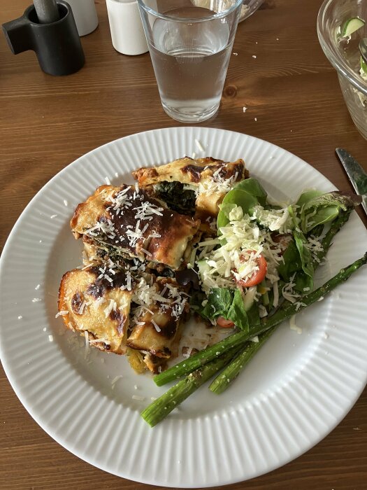 Tallrik med lasagne, grönsallad, riven ost och några grillade sparrisar. Vattenglas på ett trämöblerat matbord.