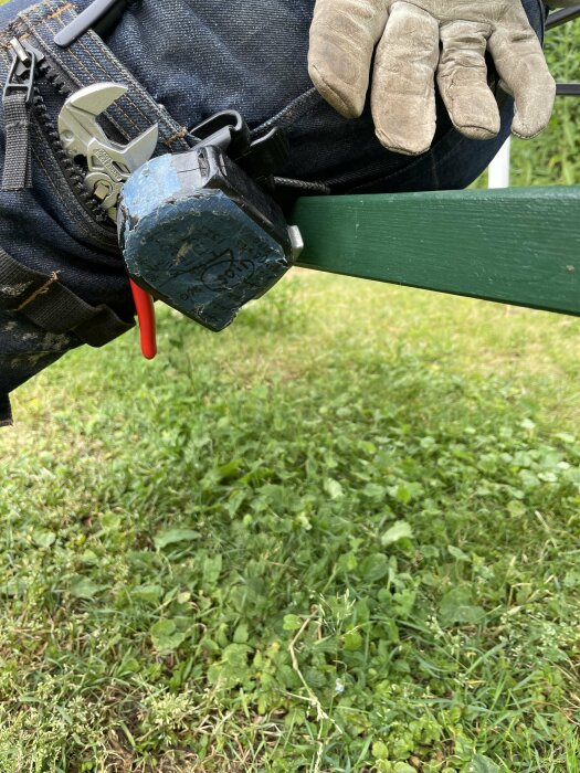 Verktygsbälte med hammare och handske hängande på grön staket; gräsmatta i bakgrunden.