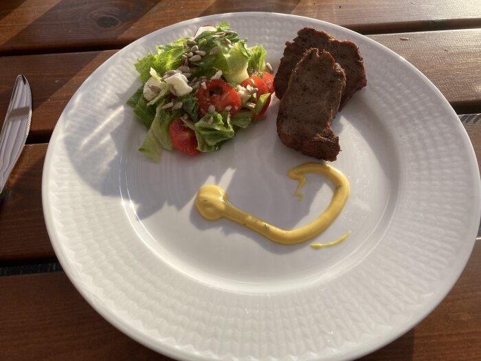 En tallrik med sallad, tomater, solrosfrön, köttbit och senapsklick på träbord utomhus. Soligt väder.