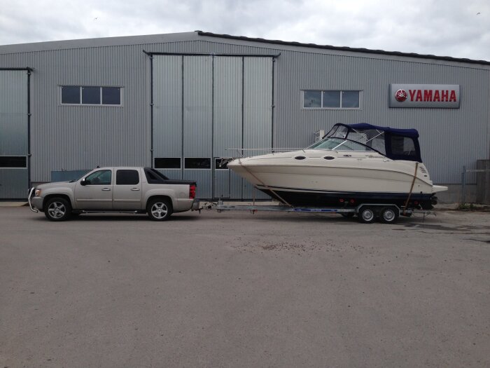 Pickup lastar en båt på trailer vid en industribyggnad med Yamaha-skylt. Grå himmel, grusmark.