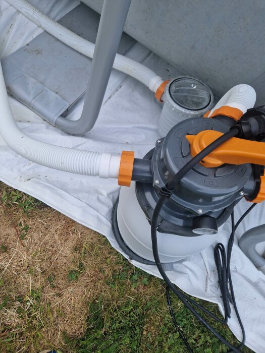 Poolpump och filter på gräs bredvid grått staket, vita och orange detaljer, sladdar och slangar.