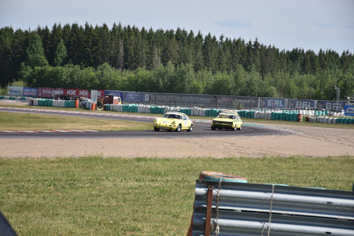 Två gula klassiska bilar tävlar på en racerbanan omgiven av skog och säkerhetsbarriärer.