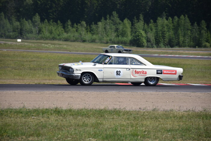 En vit vintage racingbil med startnummer 92 körs på en racerbana, omgiven av grönt landskap.