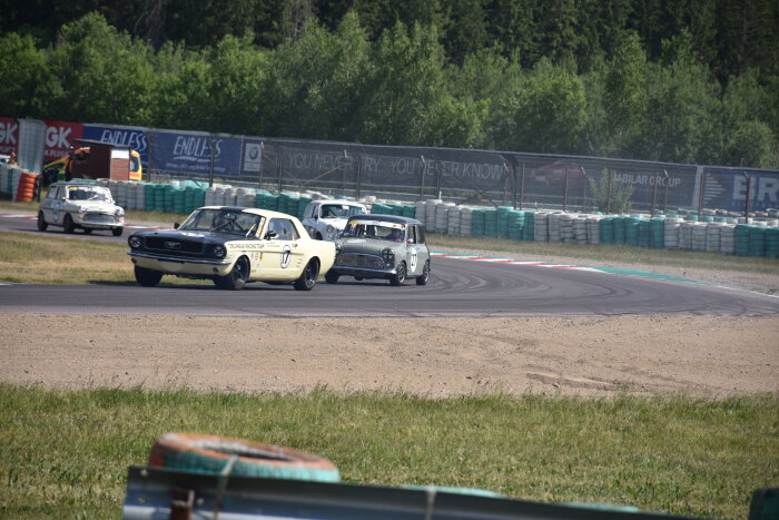 Racerbilar på bana, klassiska fordon, tävlingsmiljö, kurva, åskådare, säkerhetsstaket, motorsportevenemang, soligt, vintage.