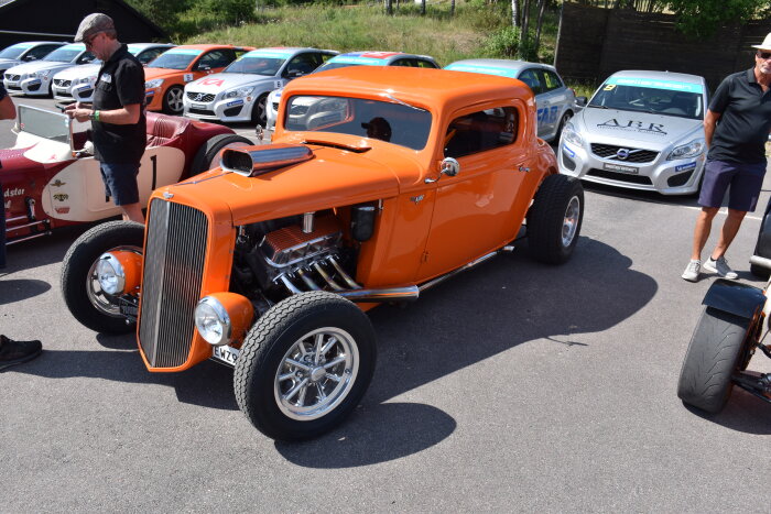 Orange hot rod-bil, utomhus bilutställning, människor tittar på, racingbilar i bakgrunden, solig dag.