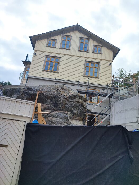 Tvåvåningshus med vit träfasad byggt på berg, omgivet av byggnadsställningar och skyddsduk, under renovering eller konstruktion.